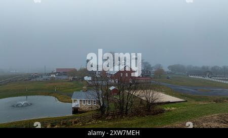 Eine rote Scheune und ein kleines Haus sitzen an einem Teich, umgeben von Gras, unter einer dicken Nebelschicht an einem bewölkten Tag. Stockfoto