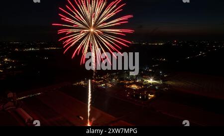 Leuchtend rotes Feuerwerk explodiert im Nachthimmel und erleuchtet Ein ländliches Messegelände mit geschäftiger Aktivität, umgeben von weitläufigen dunklen Feldern und fernen Stadtlichtern am Horizont. Stockfoto