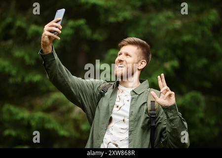 Ein bärtiger Mann lächelt, während er das Telefon in gestreckter Hand hält, inmitten eines üppigen grünen Waldes steht und einen fröhlichen Ausdruck zeigt Stockfoto