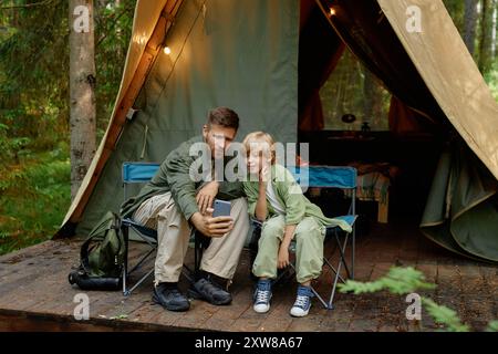 Vater und Sohn sitzen auf Stühlen vor ihrem Zelt im Wald und machen gemeinsam ein Selfie. Lächeln und Verbundenheit in natürlicher Umgebung, Zelt beleuchtet mit gemütlichen Lichtern Stockfoto