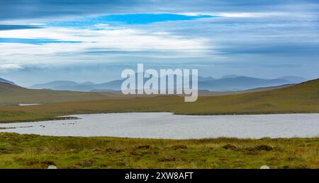 Eine ruhige Landschaft bietet ein ruhiges Gewässer, umgeben von üppigem Grün und fernen Bergen unter einem teilweise bewölkten Himmel. Stockfoto
