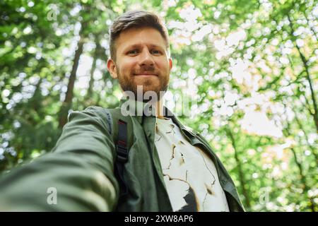 Ein lächelnder Mann macht ein Selfie in einem üppig grünen Wald, umgeben von hohen Bäumen. Sie wirken entspannt und genießen die Natur mit Sonnenlicht, das durch die Blätter filtert Stockfoto
