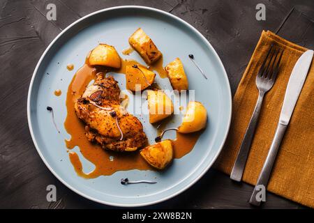 Hähnchenschenkel gebacken mit rustikalen Kartoffeln mit Ju-lie-Sauce auf einem blauen Teller auf dunklem, grauem Hintergrund und Serviette und Besteck. Hochwertige Fotos Stockfoto