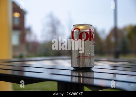 Aluminiumdose Coca-Cola Zero Sugar mit Kondenswasser – auf Metalltisch im Freien – unscharfer Hintergrund mit Bäumen und einem Gebäude in der Abenddämmerung. T Stockfoto