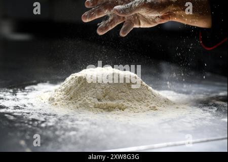 Ein erfahrener Bäcker klatscht die Hände, nachdem er Mehl auf einer Metalltheke in einer geschäftigen Bäckereiküche bestäubt hat und bereit ist, den Teig zuzubereiten Stockfoto