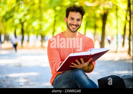 Ein junger Mann liest ein Buch in einem Park an einem sonnigen Herbsttag und konzentriert sich auf das Lernen an der Universität Stockfoto