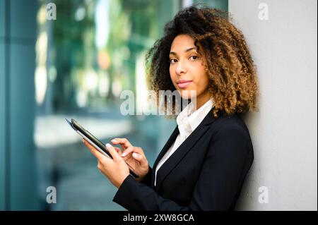 Selbstbewusste junge Geschäftsfrau, die ein digitales Tablet im modernen Büro nutzt und Erfolg und Professionalität unter Beweis stellt. Stadtbild sichtbar durch Glasfenster A Stockfoto