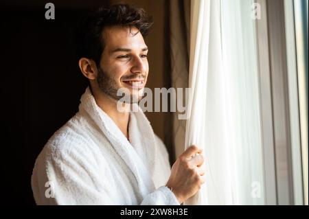 Junger Mann, der einen Bademantel trägt und vor einem Fenster in seinem Hotelzimmer schaut Stockfoto