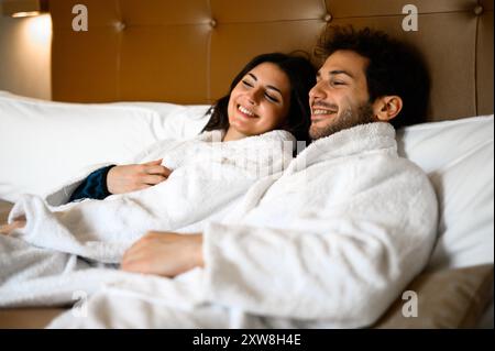 Glückliches junges Paar in weißen Bademänteln, das einen gemütlichen Moment in einem Hotelbett genießt Stockfoto