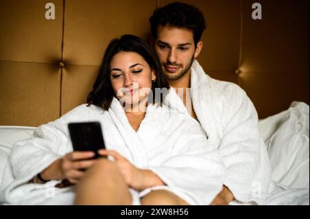 Junge Paare in Bademänteln entspannen sich im Bett, glücklich mit einem Smartphone in einem luxuriösen Hotelzimmer. Sie teilen einen Moment der Liebe und des Zusammenlebens und stöbern auf o Stockfoto