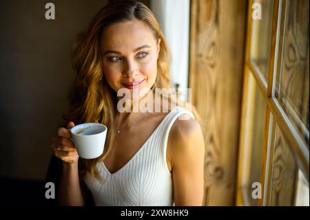 Eine junge Frau in einem weißen Kleid genießt einen friedlichen Moment in einem gemütlichen Café, blickt mit einer Tasse Kaffee aus dem Fenster und lächelt nachdenklich Stockfoto