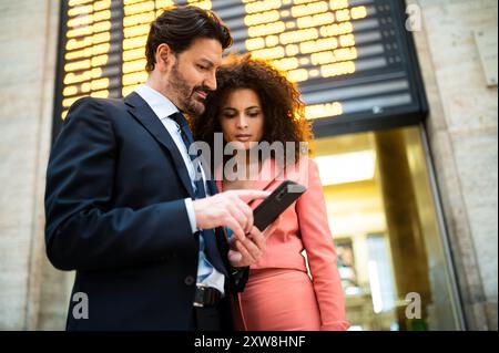 Ein Paar, das mit einem Smartphone den Fahrplan eines Bahnhofs überprüft Stockfoto