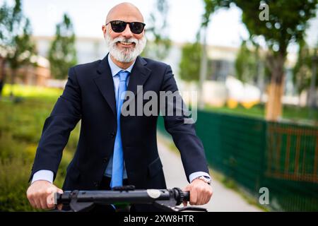 Ein reifer Geschäftsmann fährt gerne mit einem Elektroroller durch einen Stadtpark, gekleidet in einen scharfen Anzug und eine Sonnenbrille und bietet umweltfreundlichen Pendlerverkehr Stockfoto