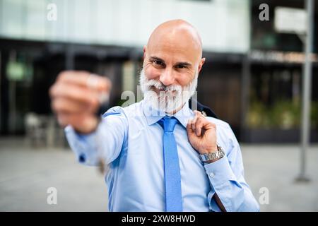 Selbstbewusster Geschäftsmann, der Schlüssel vor dem Bürogebäude hält, der Erfolg und Professionalität in scharfem blauem Anzug ausstrahlt. Urbanes Stadtbild Hintergrund hin Stockfoto