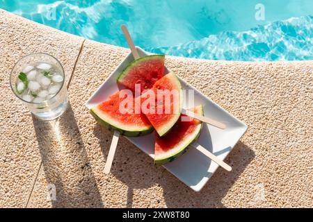 Nahaufnahme von frischen Wassermelonen-Eis auf einem Teller mit einem Glas Pfefferminzwasser am Rand eines sonnendurchfluteten Swimmingpools Stockfoto