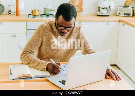 Fokussierter afroamerikanischer Mann, der von zu Hause aus arbeitet und Notizen schreibt, während er einen Laptop in einer hellen, modernen Küche benutzt. Stockfoto