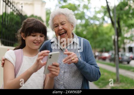 Oma holt das junge Mädchen von der Schule ab. Enkelin zeigt etwas auf dem Smartphone der Seniorengroßmutter. Stockfoto