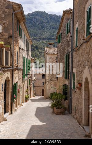 Charmante Gasse in Valldemossa, Mallorca, gesäumt von traditionellen Steinhäusern und lebendigen Topfpflanzen mit Bergen im Hintergrund. Stockfoto