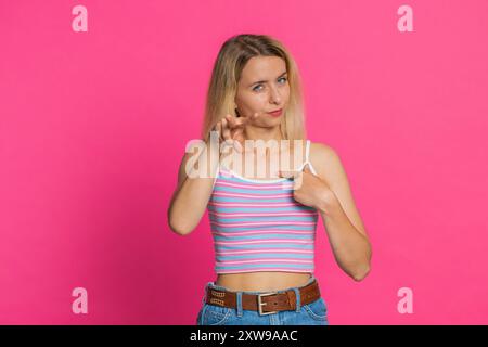 Ich beobachte dich. Selbstbewusste, aufmerksame, junge blonde Frau im Crop-Top, die auf Augen und Kamera zeigt, dass ich dich beobachte und jemanden ausspioniere. Strenge Mädchen isoliert auf rosa Hintergrund Stockfoto