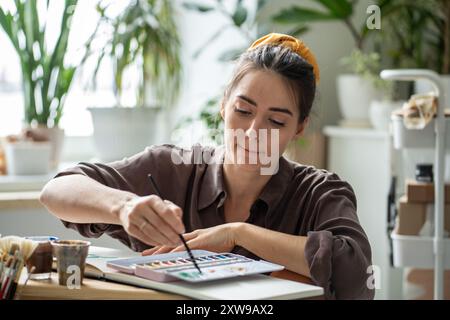 Eine fokussierte Künstlerin, die mit Aquarellen und Pinseln im Album kreiert, sitzt auf dem Boden im Kunststudio. Stockfoto