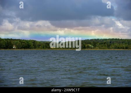 Dieses Bild zeigt eine ruhige und friedliche natürliche Landschaft mit einem ruhigen See unter einem bewölkten Himmel. Ein dezenter Regenbogen erstreckt sich über den Horizont Stockfoto