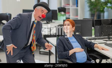 Älterer kaukasischer Mann im Clownkostüm amüsiert schwangere rothaarige Frau am Schreibtisch im Büro. Stockfoto