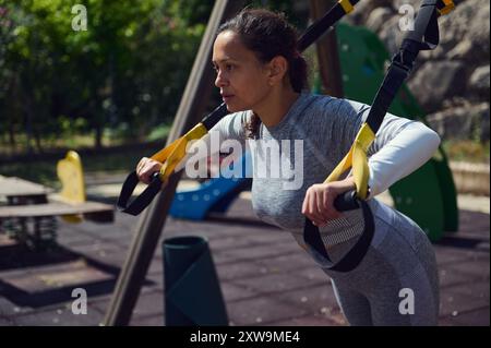 Eine entschlossene junge Frau trainiert im Freien mit Trageriemen und zeigt den Fokus und das Engagement für Fitness in einem Naturpark. Stockfoto