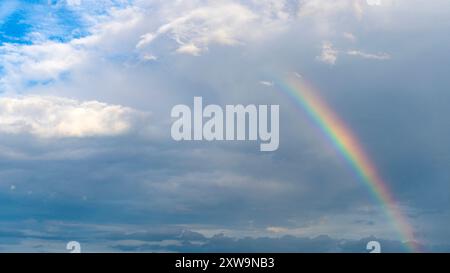 Dramatischer Sommerhimmel mit wunderschönem Regenbogen, der aus der Wolke auftaucht Stockfoto