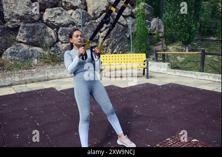 Eine Frau führt in einem Park Übungen mit Hängebändern durch, wobei der Schwerpunkt auf Fitness- und Krafttraining liegt. Der Außenbereich fügt ein natürliches Element hinzu Stockfoto