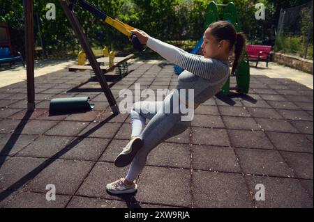 Eine Frau führt Übungen mit Aufhängungsgeräten in einem Park im Freien durch und hebt dabei Fitness, Kraft und Konzentration hervor. Stockfoto