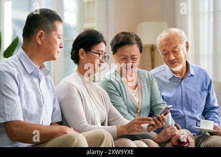 Gruppe glücklicher älterer asiatischer Leute, zwei Paare, die zu Hause auf der Familiencouch sitzen und gemeinsam auf das Handy schauen Stockfoto