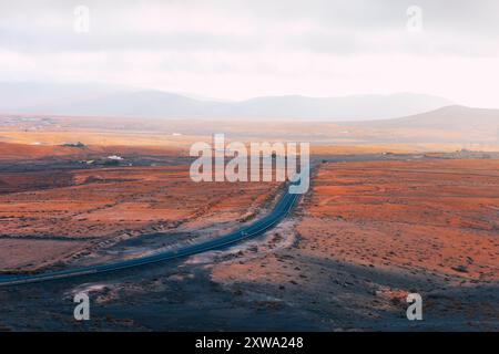 Straße in der Wüste mit Bergen in der Ferne. Gerade Straße führt durch eine weite, trockene Wüstenlandschaft auf Fuerteventura, den Kanarischen Inseln Stockfoto