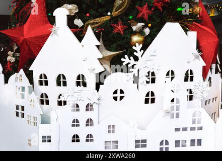 Weißer Papierausschnitt einer Stadt, die sich unter einem wunderschön geschmückten Weihnachtsbaum befindet, schafft eine festliche Weihnachtsszene. Weihnachtsbaum mit Häusern auf mir Stockfoto