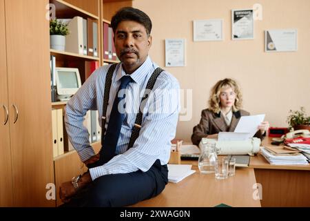 Selbstbewusster Geschäftsmann, der auf dem Schreibtisch sitzt und im Hintergrund mit einem Kollegen arbeitet, umgeben von Büroeinrichtung und Dokumenten, steht für ein professionelles Umfeld, das sich beide auf die anstehenden Aufgaben konzentriert Stockfoto