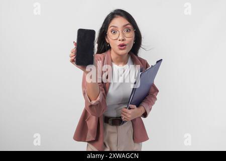 Überraschte asiatische Geschäftsfrau, die ein Dokument hält, das einen Kopierraum auf ihrem Telefonbildschirm mit schockiertem Gesicht präsentiert, isoliert von weißem Hintergrund. Stockfoto