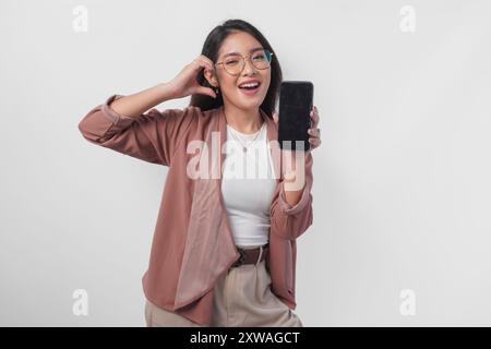 Niedliche asiatische Geschäftsfrau in Brille, die Kopierraum auf ihrem Telefon-Bildschirm präsentiert, während sie Finger-Herz-Form macht, isoliert durch weißen Hintergrund. Stockfoto