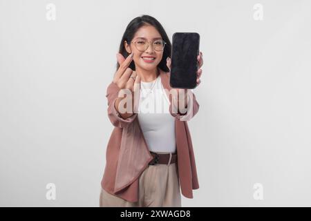 Niedliche asiatische Geschäftsfrau in Brille, die Kopierraum auf ihrem Telefon-Bildschirm präsentiert, während sie Finger-Herz-Form macht, isoliert durch weißen Hintergrund. Stockfoto