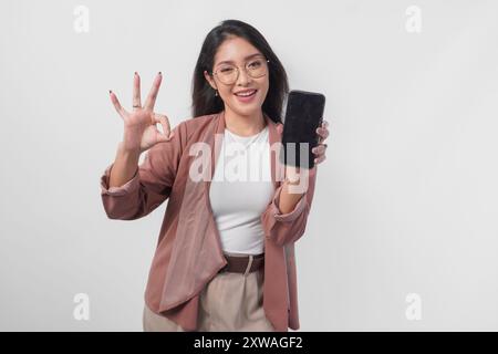 Glückliche asiatische Frau in einer Brille, die Kopierraum auf ihrem Smartphone-Bildschirm zeigt und das Zeichen „OK“ mit Gesten zeigt, isoliert von weißem Hintergrund. Stockfoto