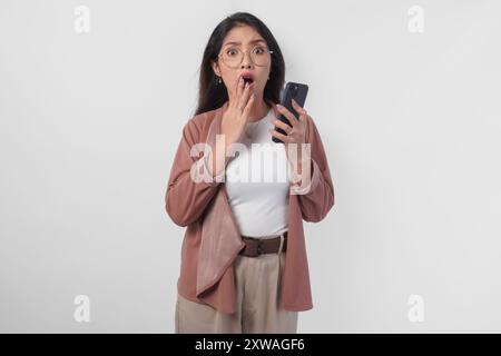 Schockierte asiatische Geschäftsfrau mit Brille, die auf ihr Smartphone zeigt, isoliert von weißem Hintergrund. Stockfoto
