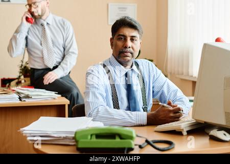 Geschäftsmann sitzt am Schreibtisch vor dem Computer, ein anderer Kollege spricht im Hintergrund telefonisch. Das Büro ist mit Papieren, Ringbuch und notwendigen Arbeitsgeräten gefüllt Stockfoto