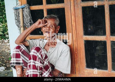 Ein nachdenklicher älterer Mann, der Saroong trägt und allein vor dem Haus sitzt und die Hände auf den Kopf gelegt hat. Stockfoto