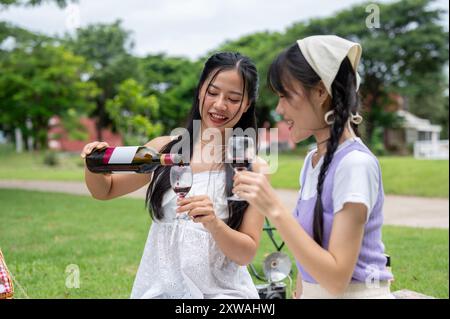 Eine schöne, glückliche asiatische Frau schenkt Rotwein für ihre Freundin, genießen Sie ein Picknick zusammen in einem grünen Park an einem hellen Tag. Glücklicher Moment, Freundschaft, Leisu Stockfoto