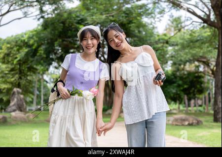 Zwei hübsche asiatische Frauen in süßen Kleidern halten Hände, schlendern durch einen Park und lächeln in die Kamera. Freundschaft, beste Freunde, LGBT Stockfoto