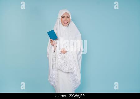 Junge asiatische muslimische Frau im Gebetskleid lächelnd und mit Pass isoliert durch blauen Hintergrund. Hajj-Konzept. Stockfoto