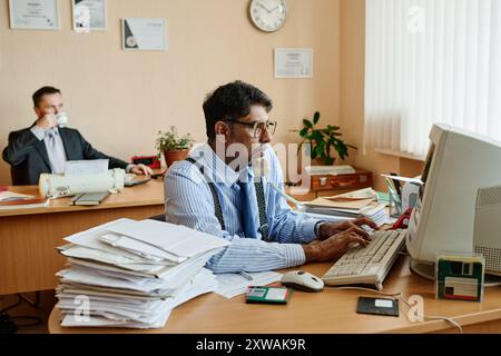 Mann mit Brille, der am Telefon spricht und am Computer mit Bürogeräten und Dokumenten auf dem Schreibtisch schreibt, während der Kollege im Hintergrund Kaffee trinkt und beobachtet Stockfoto