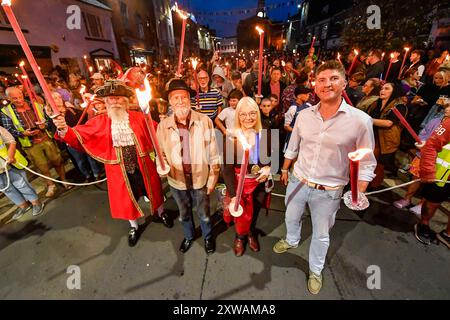 Bridport, Dorset, Großbritannien. August 2024. Hunderte von Menschen mit brennenden Fackeln nehmen an der jährlichen Bridport Carnival Fackelprozession von der Bridport Town Hall nach East Beach in der West Bay in Dorset Teil. Die Two Mile-Prozession wurde von Bridport Town Crier John Collingwood, Cllr Dave Rickard, Bridport Mayor Anne Rickard und West Dorset Parlamentsabgeordneten Edward Morello angeführt. Bildnachweis: Graham Hunt/Alamy Live News Stockfoto