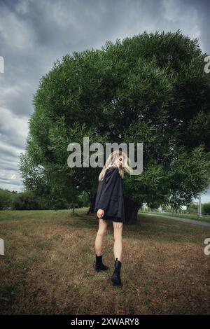 Eine junge Frau in schwarzer Jacke und Rock geht von einem großen Baum in einem Park weg. Der Himmel ist bewölkt und das Gras ist braun. Stockfoto