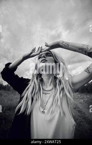 Eine Frau mit langen blonden Dreadlocks und mehreren Halsketten steht auf einem Feld und blickt auf den bewölkten Himmel. Ihre Hände sind hoch und schützen ihre Augen Stockfoto