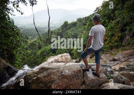 Ein Mann mit Hut steht auf einer felsigen Klippe mit Blick auf einen kaskadierenden Wasserfall, mit einem atemberaubenden Blick auf eine üppige, grüne, bewaldete Landschaft im Hinterland Stockfoto