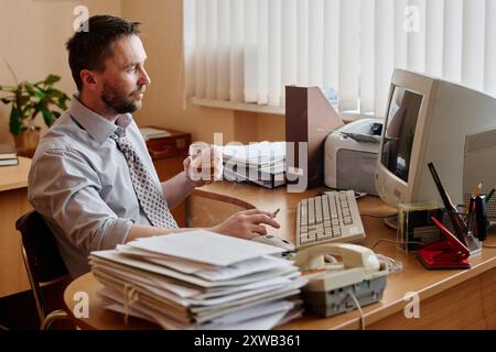 Kaukasischer Mann sitzt am Holzschreibtisch mit einem Glas Getränk in der Hand, umgeben von Bürobedarf und Technologie in hellem Büroumfeld Stockfoto